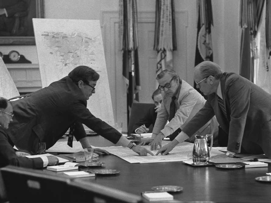 Men discussing around a table pointing to a piece of paper on the table between them.