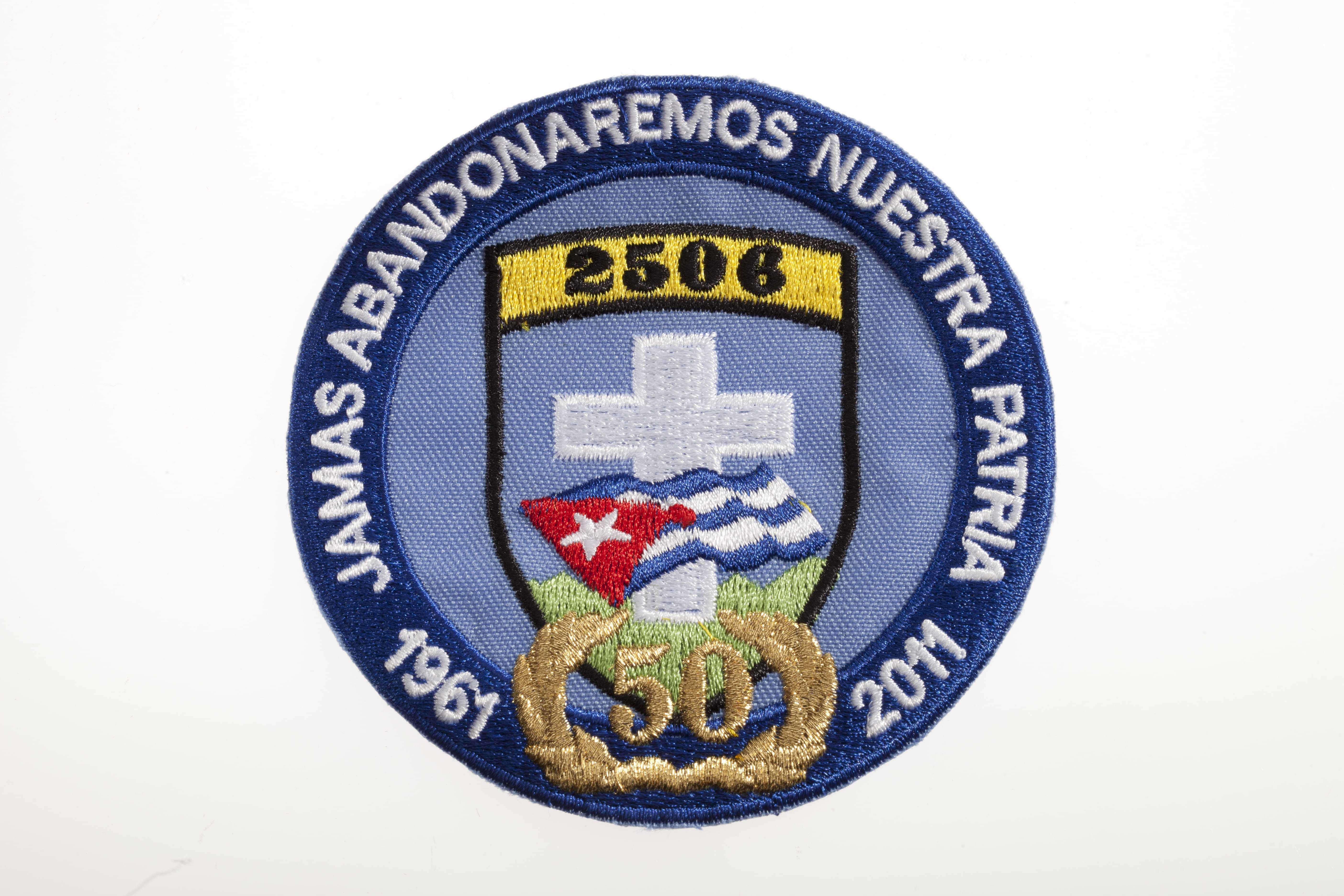 A blue circular patch with a Cuban flag and a cross in the center. The dates 1961 and 2011 are placed on opposite sides of the number 50.