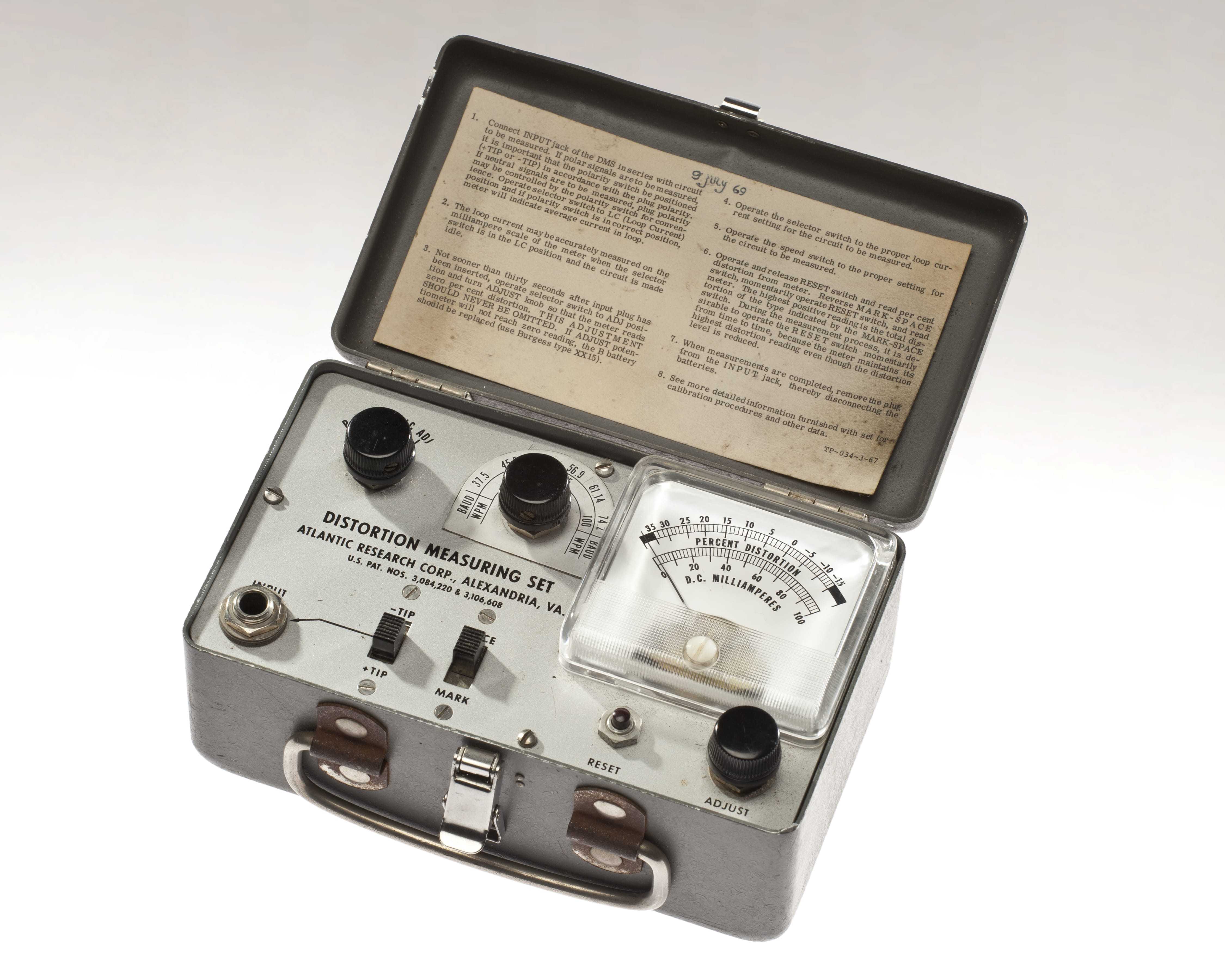 An open metal box containing instructions for its use, various switches and dials, and a distortion level meter
