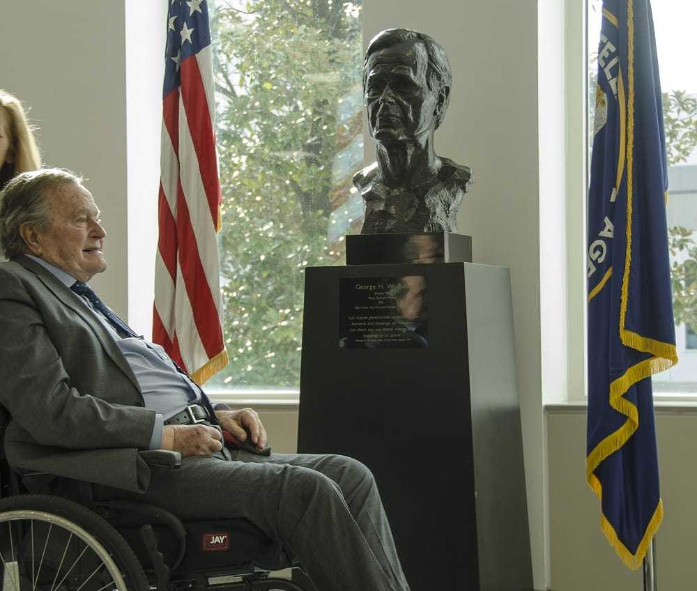 Bush, in a wheelchair, smiling at his bust statue sitting between the American glad and the CIA flag.