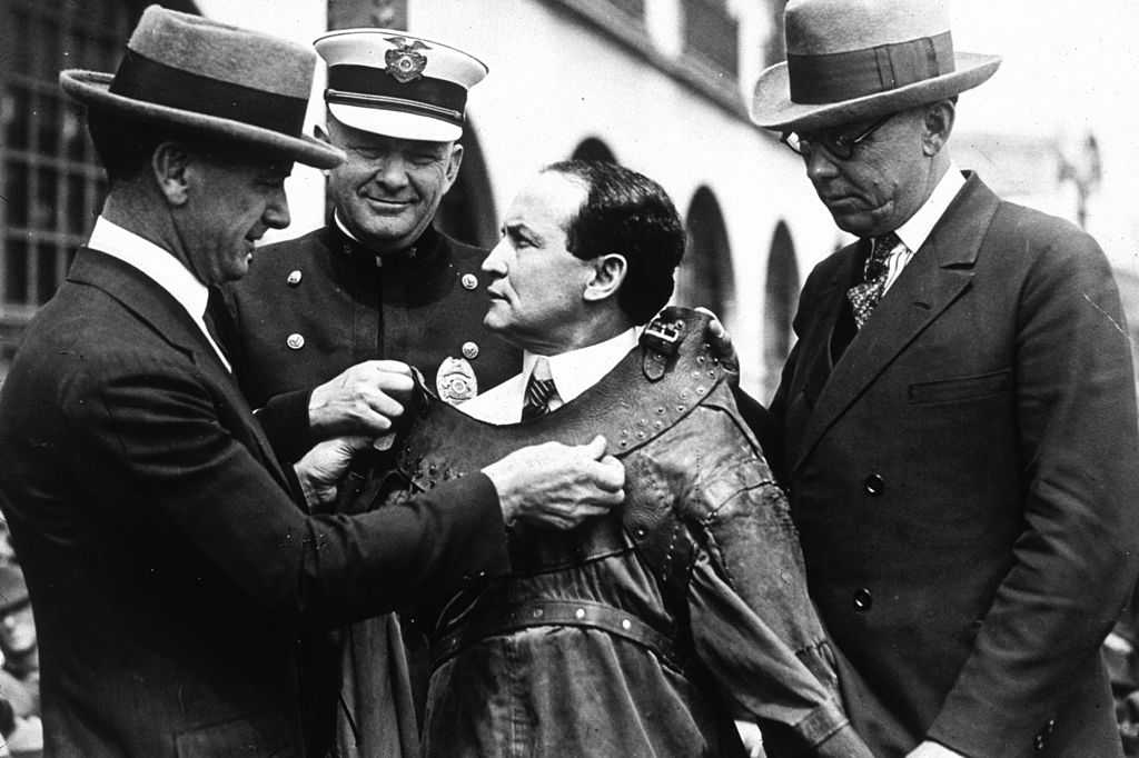 Old black and white photograph of Harry Houdini surrounding by police officers who are fitting him into a straight jacket for a performance.