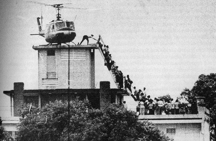 A black and white photograph of a helicopter perched atop a small building. A long line of evacuees are climbing a ladder to reach the helicopter.