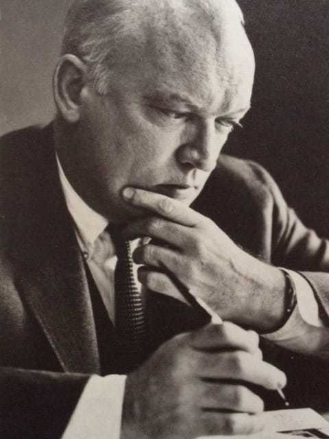 A black and white photograph of a pensive James with his left hand on his chin and a pen in his right hand.