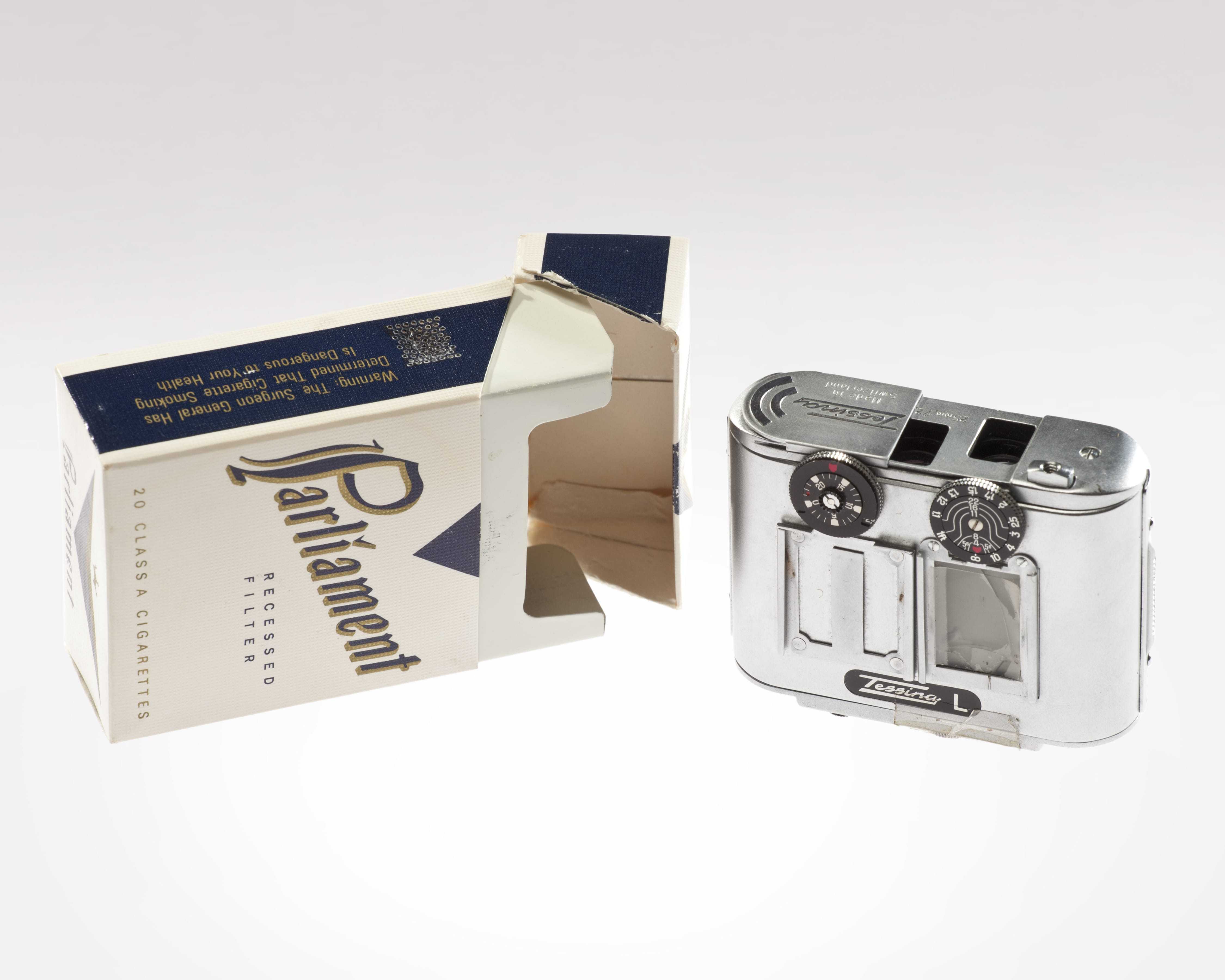 An empty box of Parliament cigarettes next to a smaller film camera with a silver metal exterior