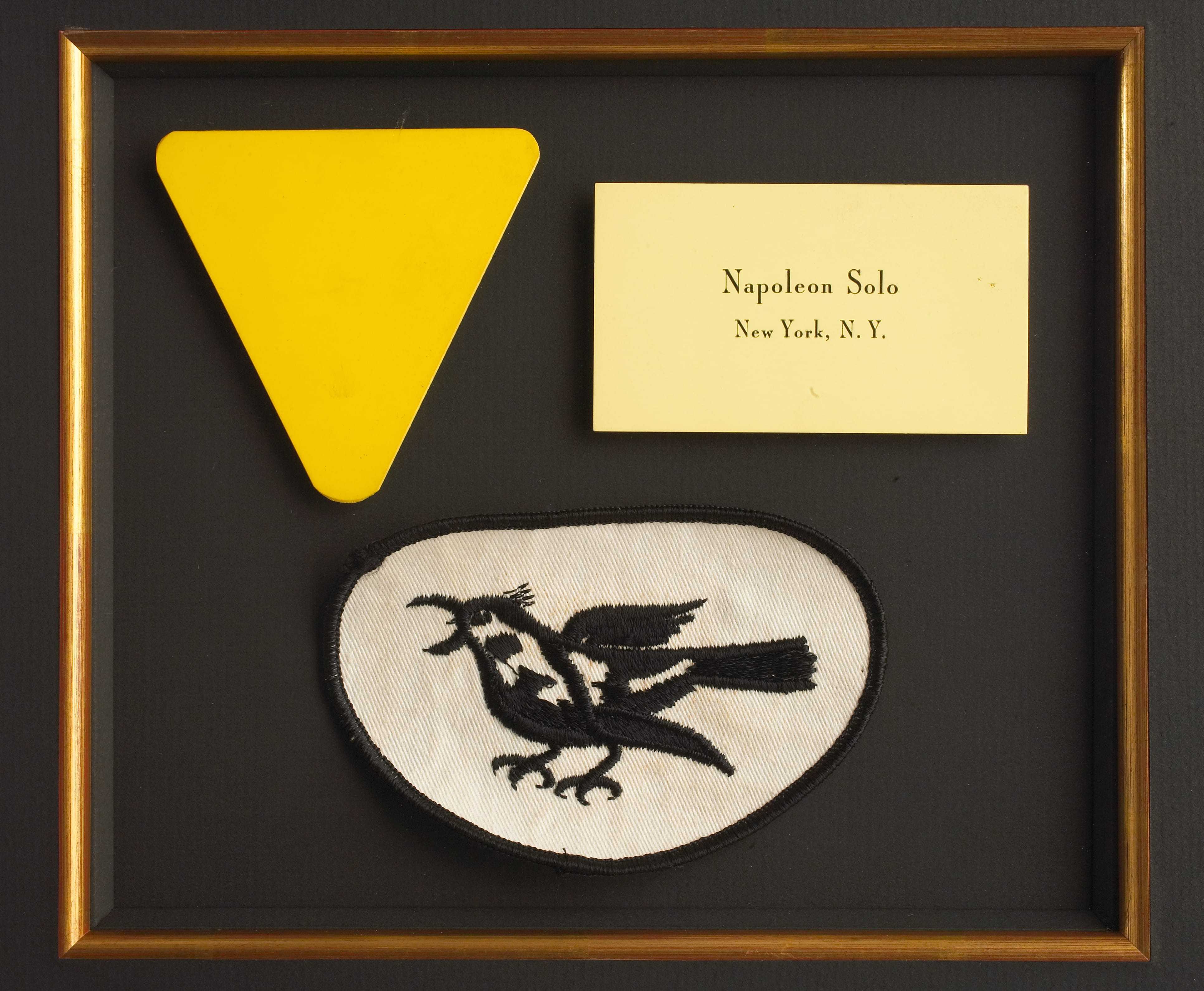 A yellow security badge, a patch with a bird insignia, and an business card for Napoleon Solo