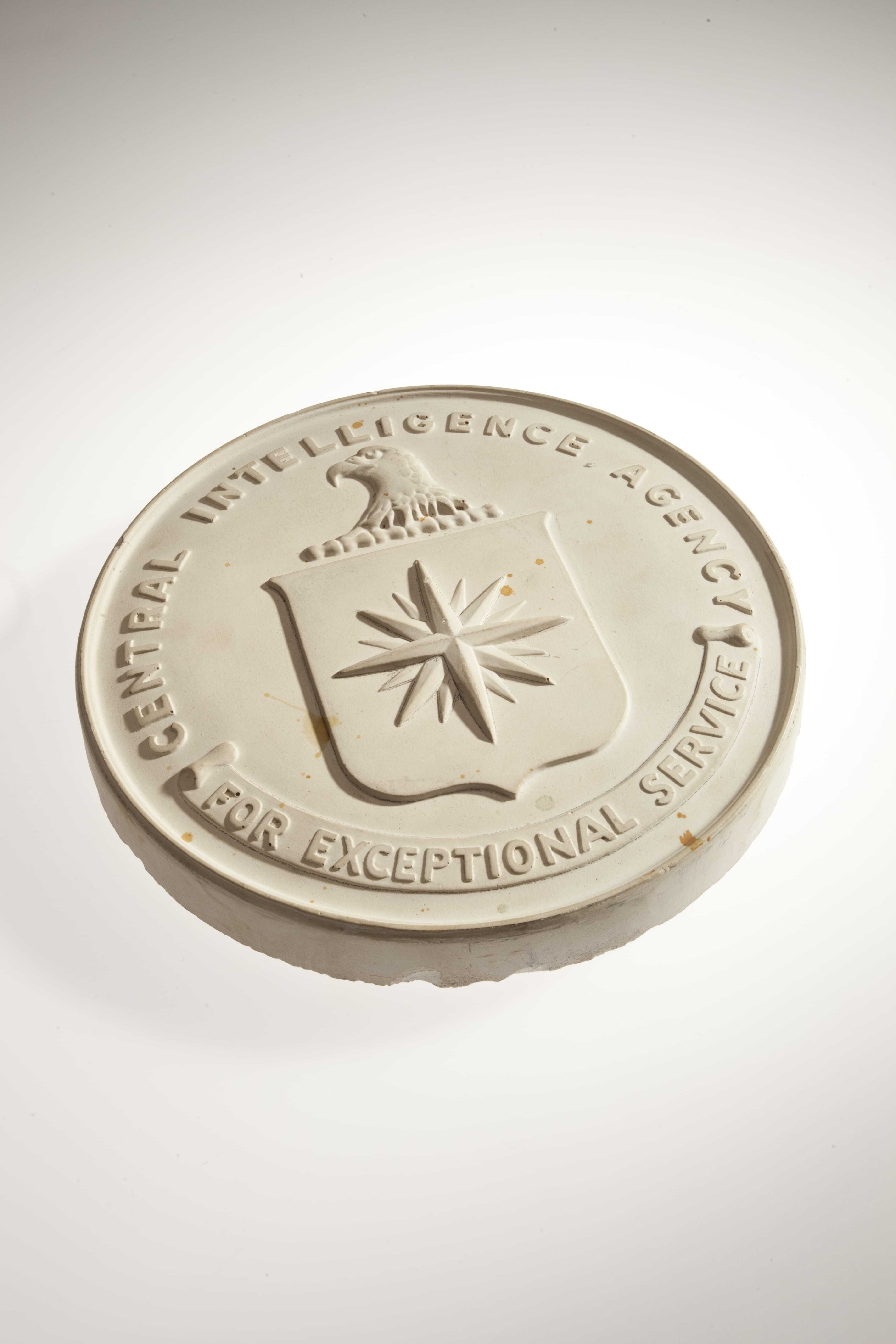 An ivory colored plaster cast of the CIA Seal with the CIA's motto "For Exceptional Service" inscribed at the bottom.