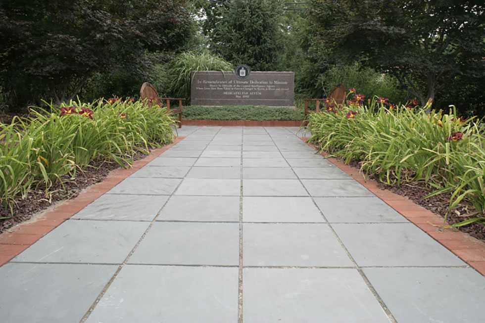 A brown, granite memorial wall with an engraved dedication, surrounded by a stone walkway, landscaped greenery, and benches.