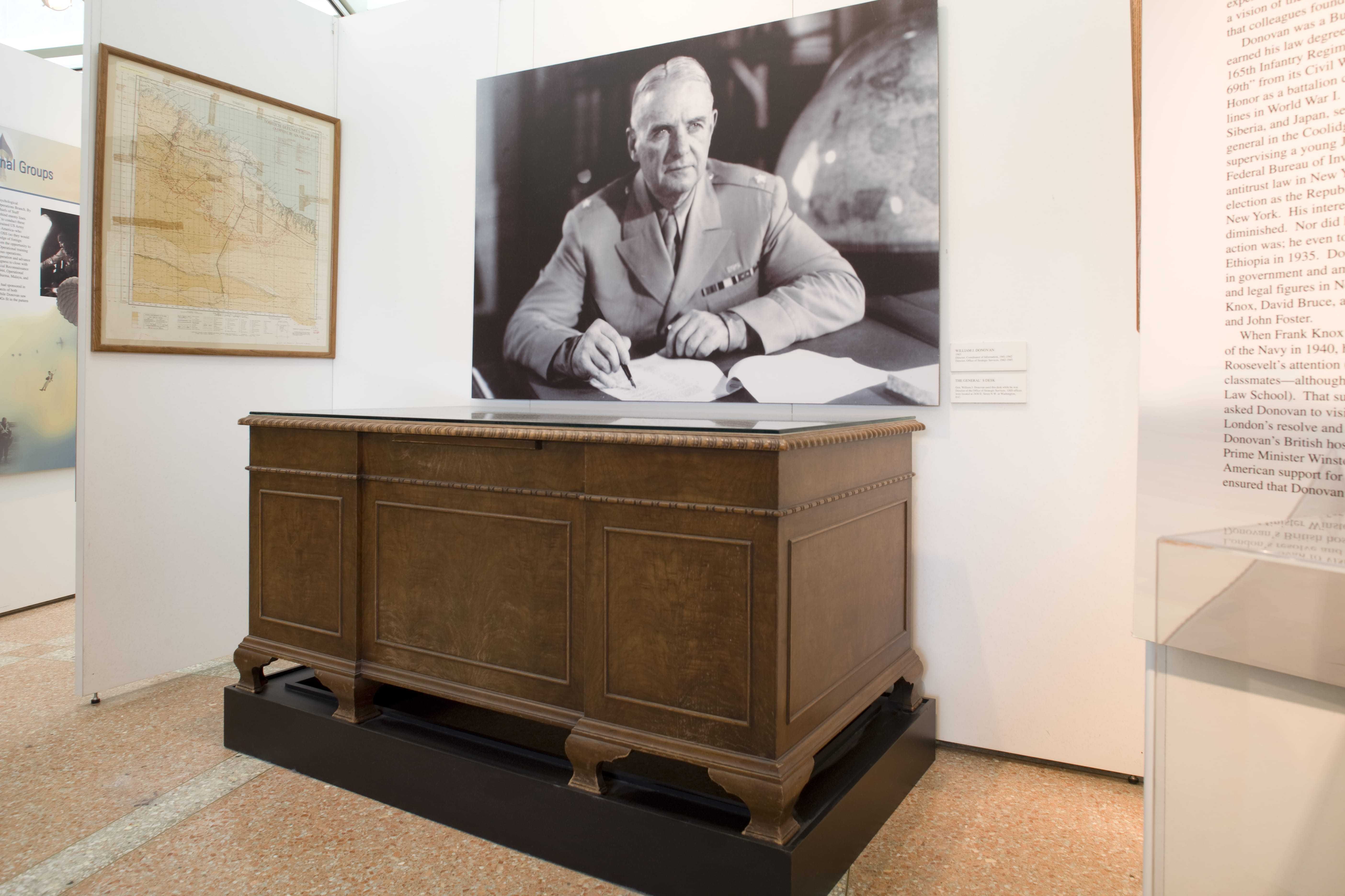 A large wooden desk with a glass top in front of a poster of William J Donovan