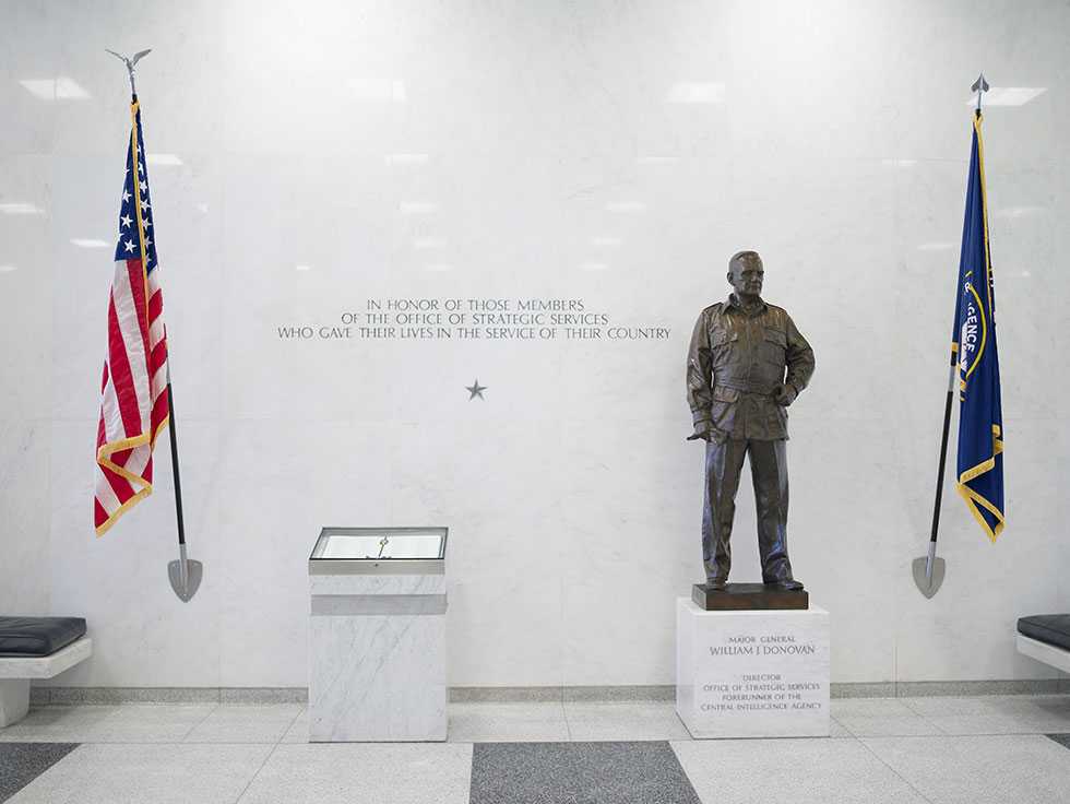 The American flag, a book listing the names of fallen OSS officers encased in a display case, and a bronze-colored statue of Maj. Gen. William J. Donovan amidst a white wall with an inscription engraved into it.