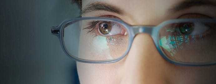 A close up of a woman's eyes, with computer screens reflected off her glasses.