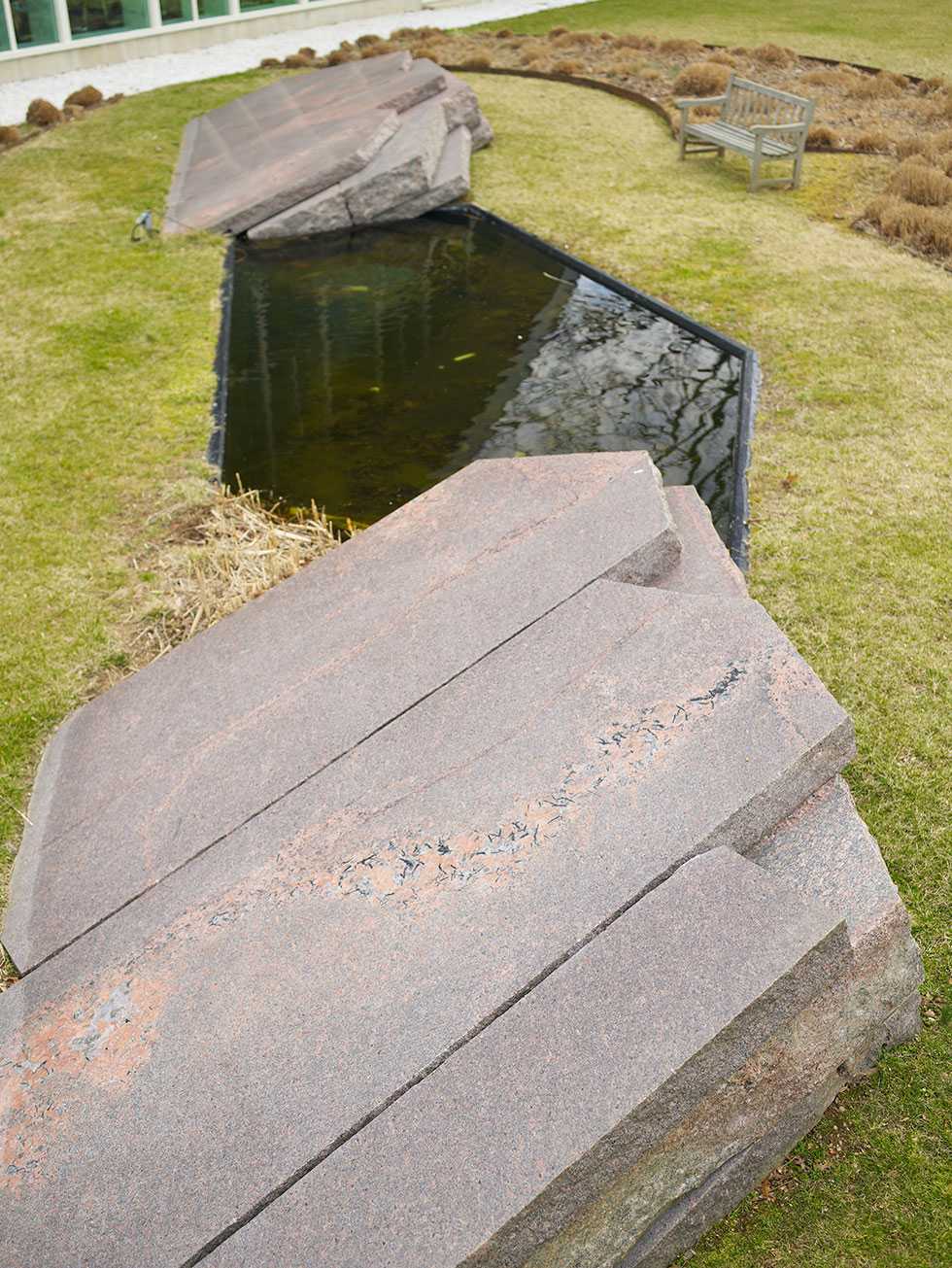 A black hexagonal pond in a patch of grass with large slated stones, a bench and shrubbery on a grassy area.