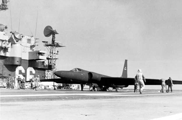 A black and white image of an an aircraft on the deck of a naval ship with three people standing near it.