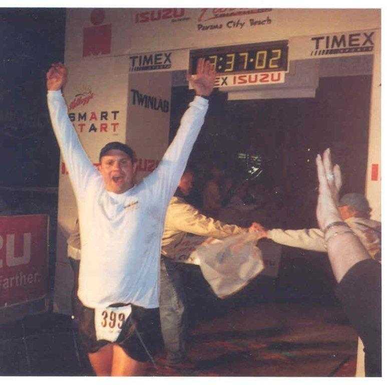 A color photograph of Gregg raising his arms with excitement after completing an Ironman.
