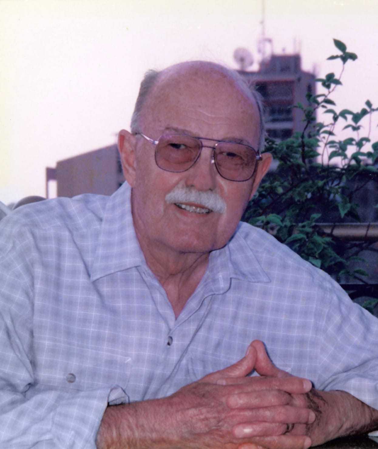 A color portrait photograph of an older and balding LaGueux with a thick white mustache, a checkered dress shirt unbuttoned at the top, and large stylish sunglasses.