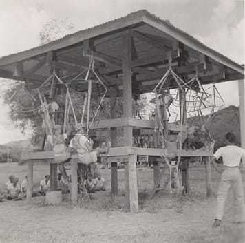 Soldiers sitting in parachute harnesses suspended from wooden beams.