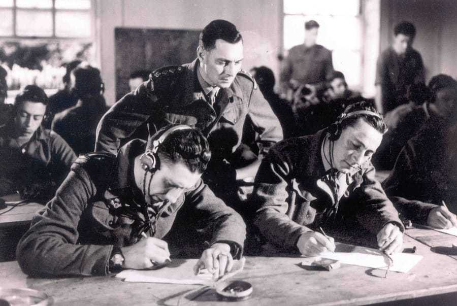 Two students taking notes at a desk with a man peering over them from behind.