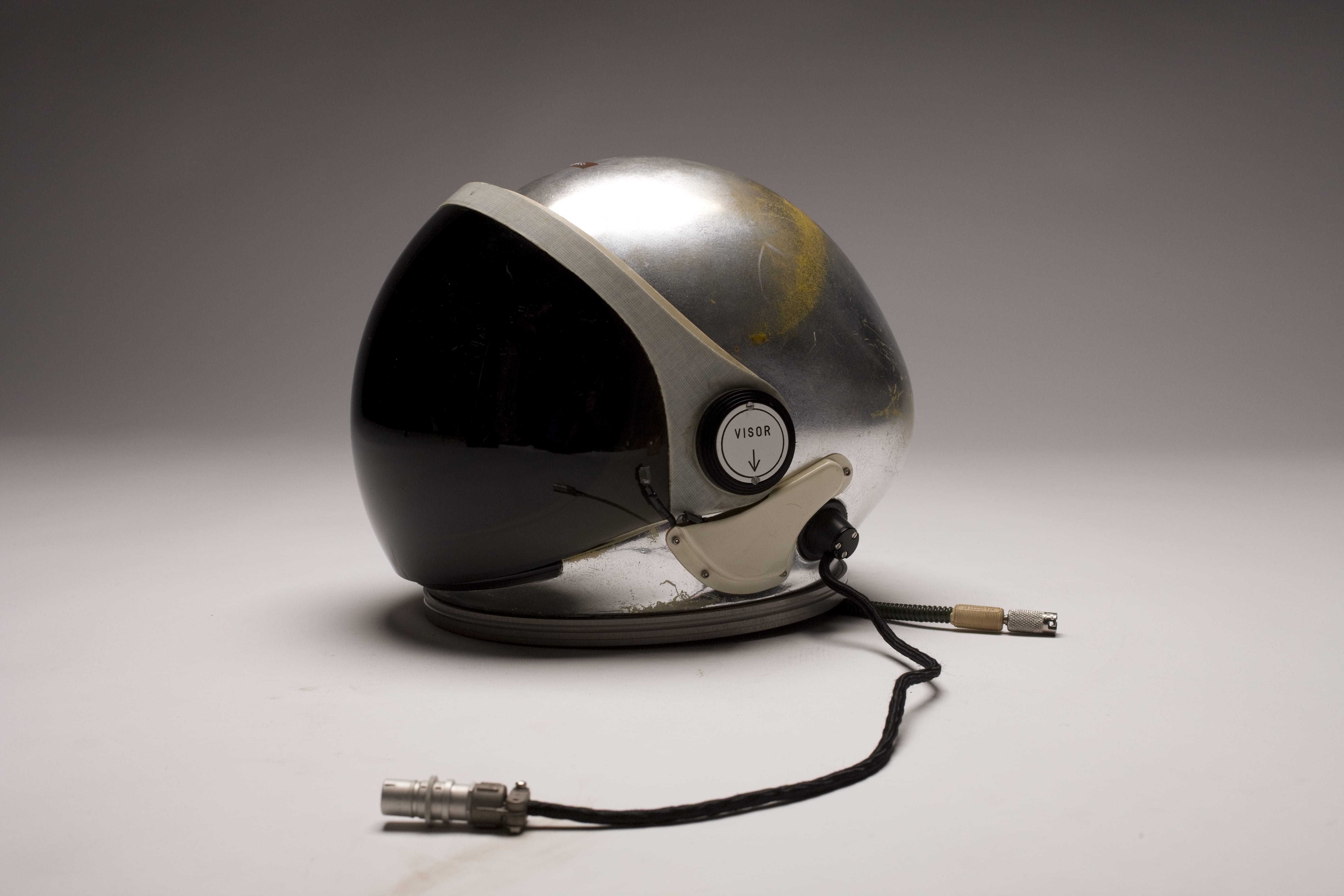 A silver A-12 pilot's helmet with a large visor and cords to connect to the suit