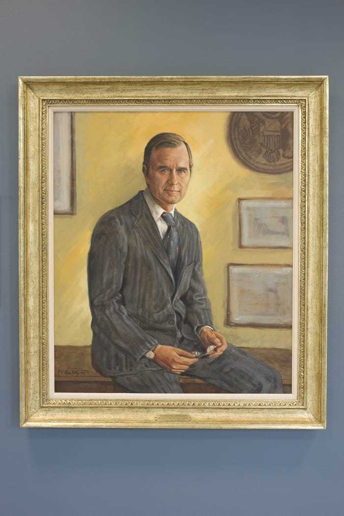 A painting of Bush sitting on a desk, with his hands laying on his lap, in a gold frame hanging on a blue wall.
