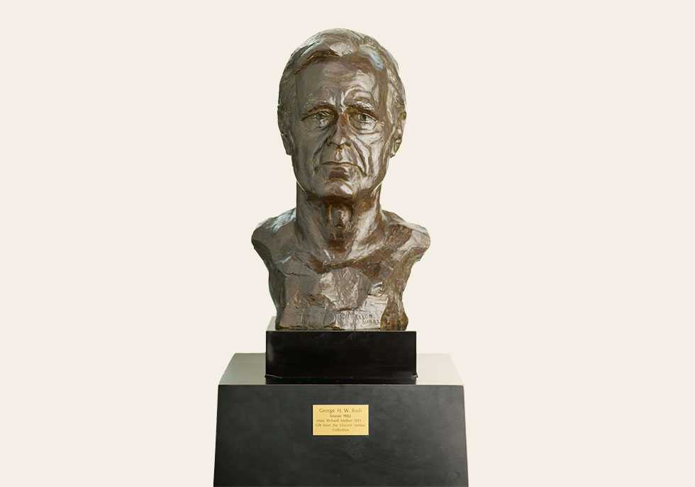 A brown, stone sculpture of a bust on a black pedestal with a gold commemorative plaque.