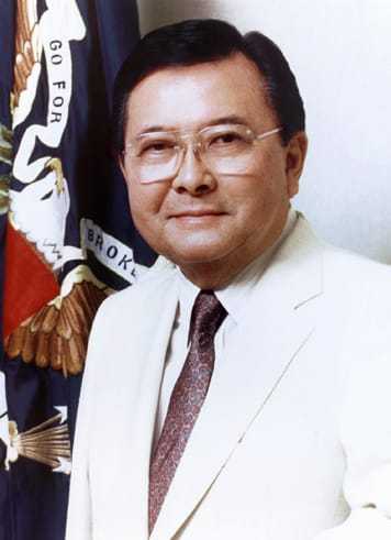 A man with dark hair and glasses grinning in front of a flag in a white suit coat.