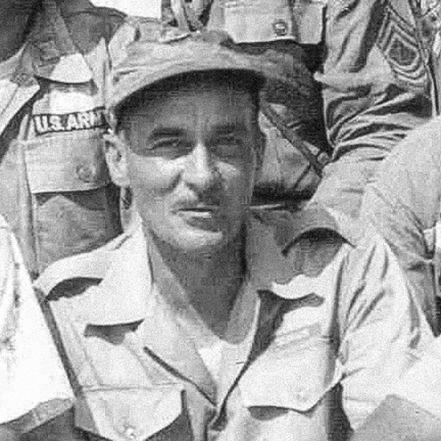 A black and white close up of Jennings Benson smirking at the camera in his army uniform surrounded by other soldiers in uniform.