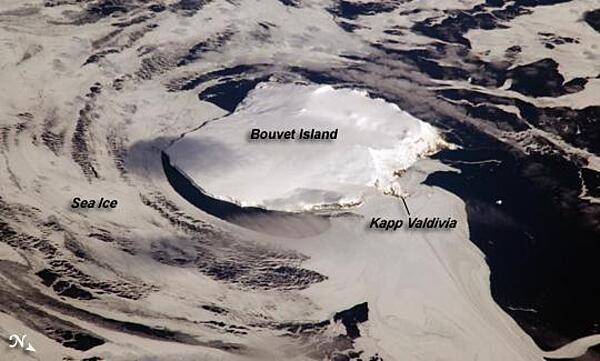 Bouvet Island is one of the most remote islands in the world; Antarctica, over 1,600 km (1,000 mi) to the south, is the nearest land mass. Located near the junction of three tectonic plates, the island is mostly formed from a shield volcano that is almost entirely covered by glaciers. The prominent Kapp (Cape) Valdivia on the northern coastline is a peninsula formed by a lava dome. It is only along the steep cliffs of the coastline that the underlying dark volcanic rock is visible against the white snow and ice blanketing the island. Photo courtesy of NASA.