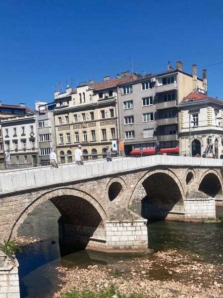 The Latin Bridge is the oldest of over a dozen bridges crossing the Miljacka River in Sarajevo. The original wooden bridge dates to around 1541 but was replaced by a stone structure in 1565. A 1791 flood damaged the bridge, and it was rebuilt around 1798. Constructed while Bosnia was under the Ottoman Empire, the bridge acquired its name from the fact that it connected with the Catholic quarter of the city (“Latinluk”) on the right bank of the river. The stone and gypsum bridge has four arches and sits on three pillars that have two relieving openings or “eyes” above them. The distinctive “eyes” also appear on the Sarajevo coat of arms. The Latin Bridge acquired a place in history as the spot where World War I began. On 28 June 1914 at the foot of the bridge, a Serb nationalist, Gavrilo Princip, shot and killed Franz Ferdinand, heir presumptive to the Austro-Hungarian throne, and his wife Sophie sparking the conflagration.