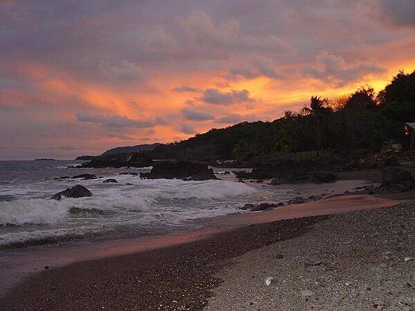 Fiery clouds hover over a darkened jungle coastline at sunset.