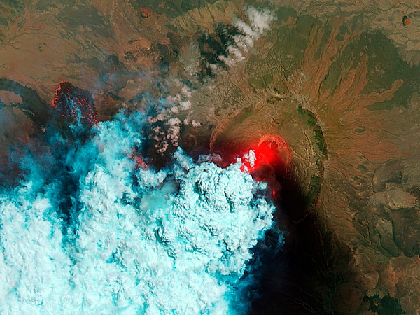 Satellite view of the Nabro volcano in Eritrea, taken on 24 June 2011. The bright red areas indicate hot surfaces. Hot volcanic ash glows above the vent, located in the center of Nabro’s caldera. To the west of the vent, portions of an active lava flow are also hot. The speckled pattern on upstream portions of the flow are likely due to the cool, hardened crust splitting and exposing fluid lava as the flow advances. The bulbous blue-white cloud near the vent is likely composed largely of escaping water vapor that condensed as the plume rose and cooled. The wispy, cyan clouds above the lava flow are evidence of degassing from the lava. Image courtesy of NASA.