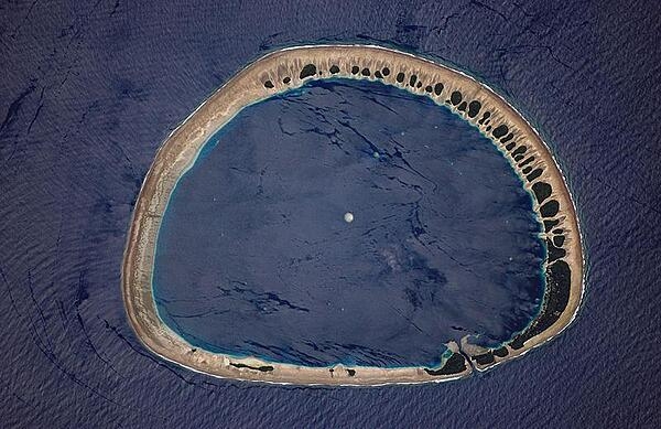 Classically shaped Nukuoro Atoll is one of 607 islands that make up the Federated States of Micronesia. It is also a municipality of the state of Pohnpei, Federated States of Micronesia. The atoll has a population of only several hundred people. The total area, including the lagoon, is 40 sq km, with a land area of only 1.7 sq km, which is divided among more than 40 islets that lie on the northern, eastern, and southern sides of the lagoon. By far the largest islet is Nukuoro islet in the southeast, which is the center of population and the capital of the municipality. Vegetation is most prominent on the northeast and east portions of the atoll that face the dominant Easterly winds.  Swells driven by these winds can be seen approaching from the east-southeast (right) and wrapping around the atoll to produce an interference pattern on the downwind side. Water in the lagoon is calm in comparison. A coral head appears dead center of the photo. Image courtesy of NASA.