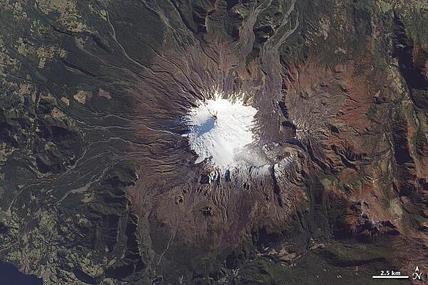 Near the southern tip of South America in Chile, the most active of three volcanoes is the westernmost, Volcan Villarrica, pictured in this photo-like satellite image. The 2,582 m (9,357 ft) stratovolcano is mantled by a 30 sq km (10 sq mi) glacier field, most of it south and east of the summit in a basin made by a caldera depression. To the east and northeast, the glacier is covered by ash and other volcanic debris, giving it a rumpled, brown look. The western slopes are streaked with gray-brown gullies, the paths of lava and mudflows (lahars). Image courtesy of NASA.