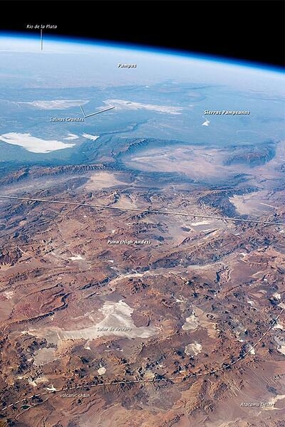 This panorama looking southeast across the South American continent was taken from the International Space Station almost directly over the Atacama Desert near Chile's Pacific coast. The high plains (3000-5000 m, 13,000-19,000 ft) of the Andes Mountains, also known as the Puna, appear in the foreground, with a line of young volcanoes (dashed line) facing the much lower Atacama Desert (1000-2000 m elevation). Several salt-crusted dry lakes occupy the basins between major thrust faults in the Puna. Salar de Arizaro (foreground) is the largest of the dry lakes in this view. The Atlantic Ocean coastline is dimly visible at the top left. Near image center is the transition between two distinct geological zones, the Puna and the Sierras Pampeanas. The color change from reds and browns in the foreground to blues and greens in the upper part of the image reflects the major climatic regions: the deserts of the Atacama and Puna versus the grassy plains of central Argentina. Image courtesy of NASA.