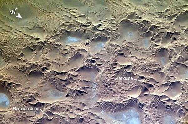 The Issaouane Erg (sand sea) is located in eastern Algeria. Ergs are vast areas of moving sand with little to no vegetation cover. The most common landforms in the image are star dunes and barchan (or crescent) dunes. Star dunes are formed when sand is transported from variable wind directions, whereas barchan dunes form in a single dominant wind regime. Occasional precipitation fills basins formed by the dunes; as the water evaporates, salt deposits are left behind which appear as bluish-white areas. Image courtesy of NASA.