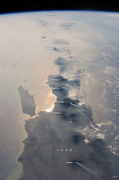 This photograph, taken from the International Space Station in 2015, looks eastward: Java is in the foreground, Bali and Lombok are near the center, and smaller islands trail off toward the horizon. The brightest reflection of the Sun off the sea surface silhouettes Surabaya, Indonesia’s second-largest city of almost 3 million. Against this background of glint as well as regional smoke, a line of volcanoes appears in sharp detail. Volcanoes are the backbone of the islands, which have been formed by the collision of the Australian tectonic plate (right) with the Asian plate (left). The name of each volcano appears in italics. White plumes - each about 80 km (50  mi) long - show that at least six volcanoes appeared to be emitting steam and smoke. The plumes are strikingly parallel, aligned with winds from the northeast. Image courtesy of NASA.